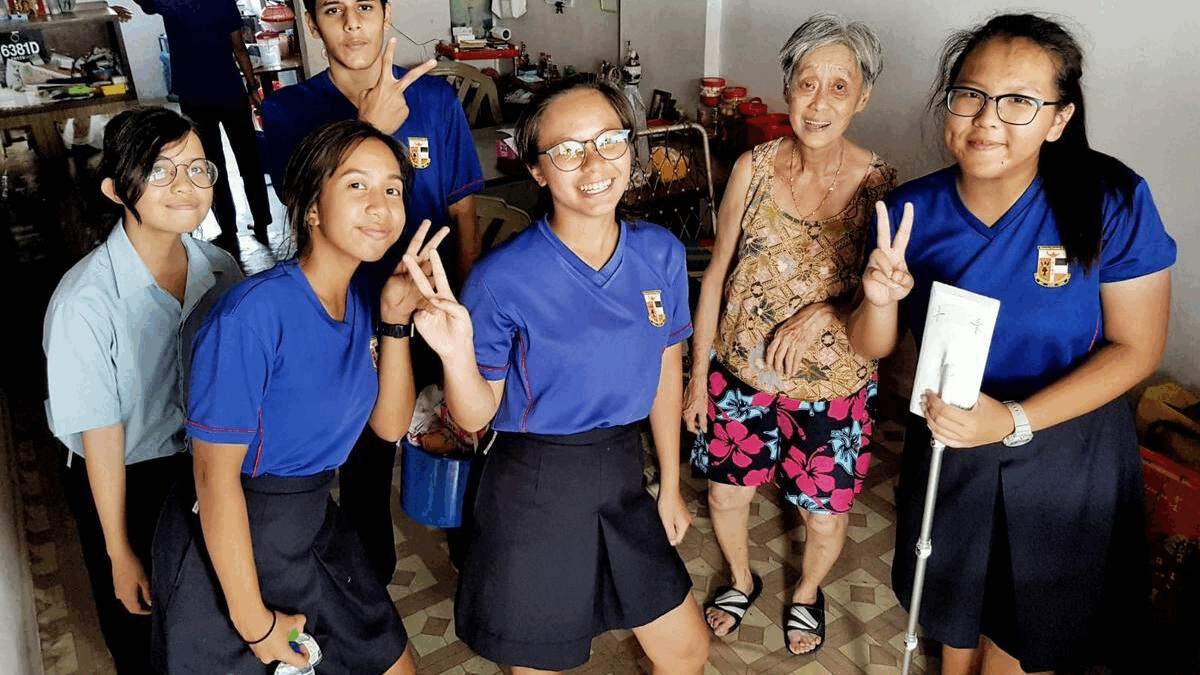 Five students in blue uniforms pose with an elderly woman indoors, several making peace signs and smiling.